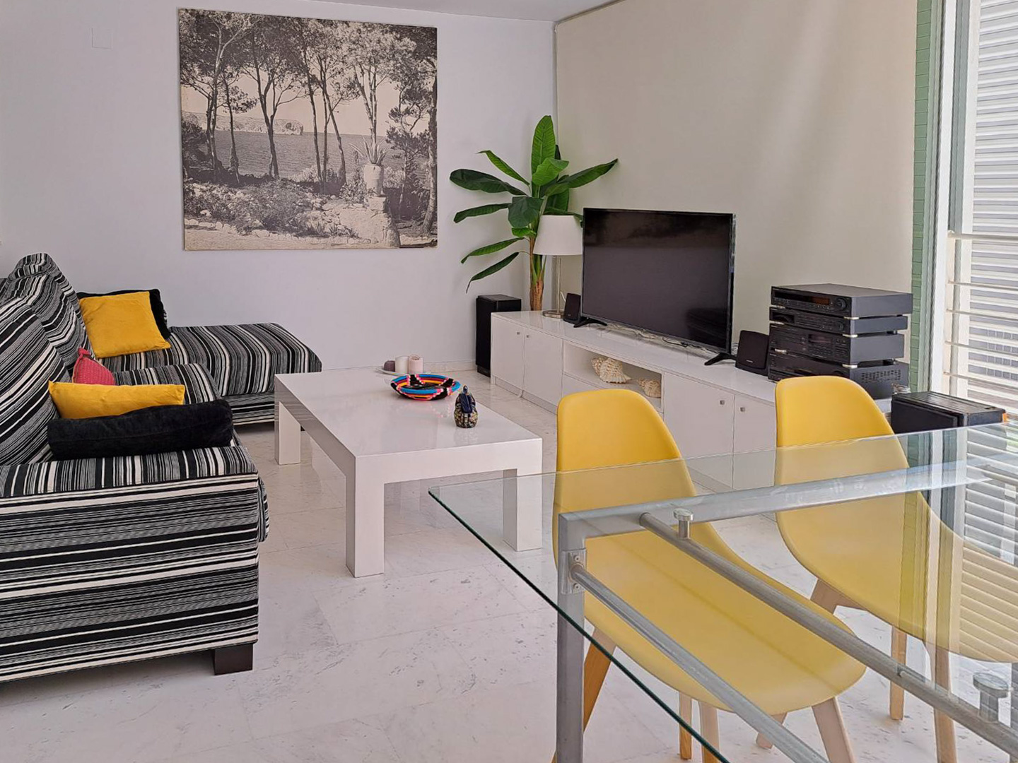 Espace de vie avec sol en marbre, canapé à rayures et coin repas composé d'une table en verre et chaises jaunes. Un meuble TV blanc complète cet intérieur lumineux.