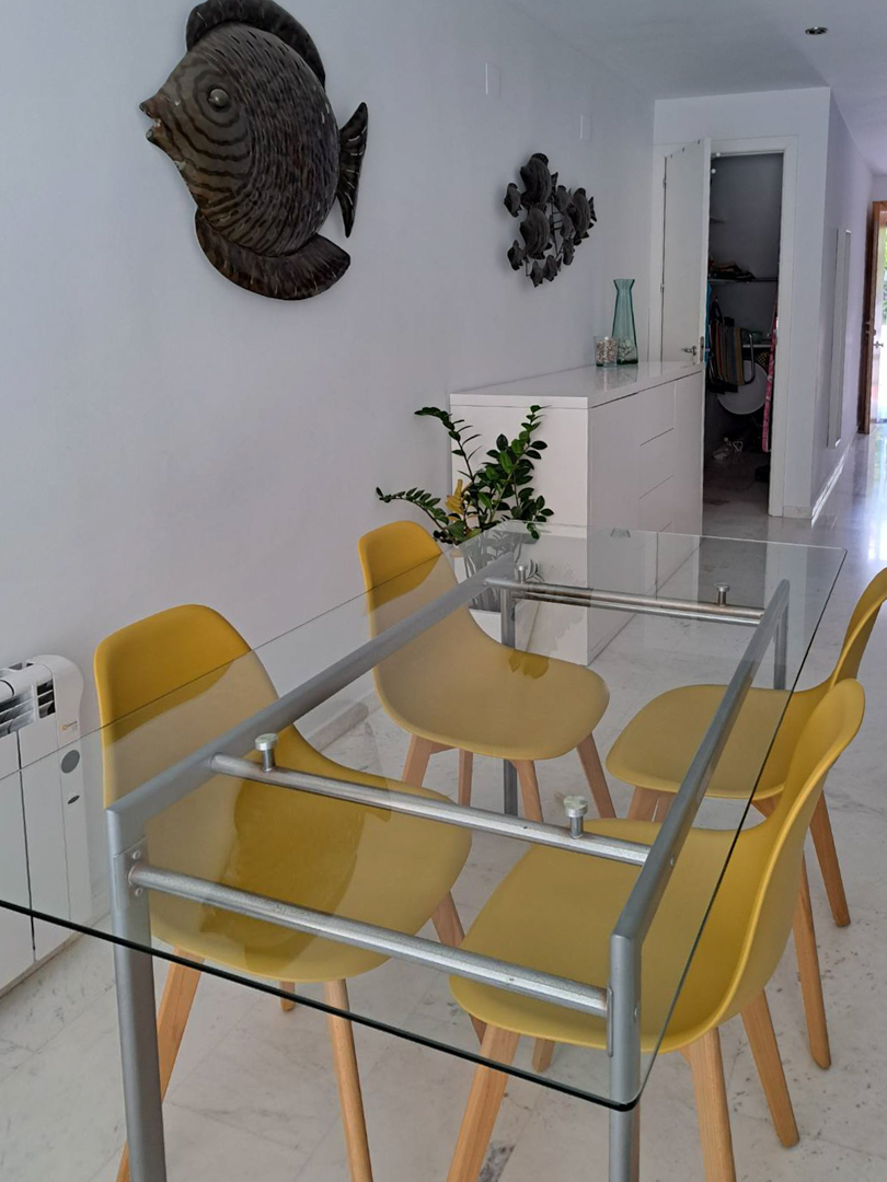Espace repas avec table en verre et chaises jaunes sur sol en marbre. La pièce dispose d'un buffet blanc, d'un radiateur électrique et d'une décoration murale marine.