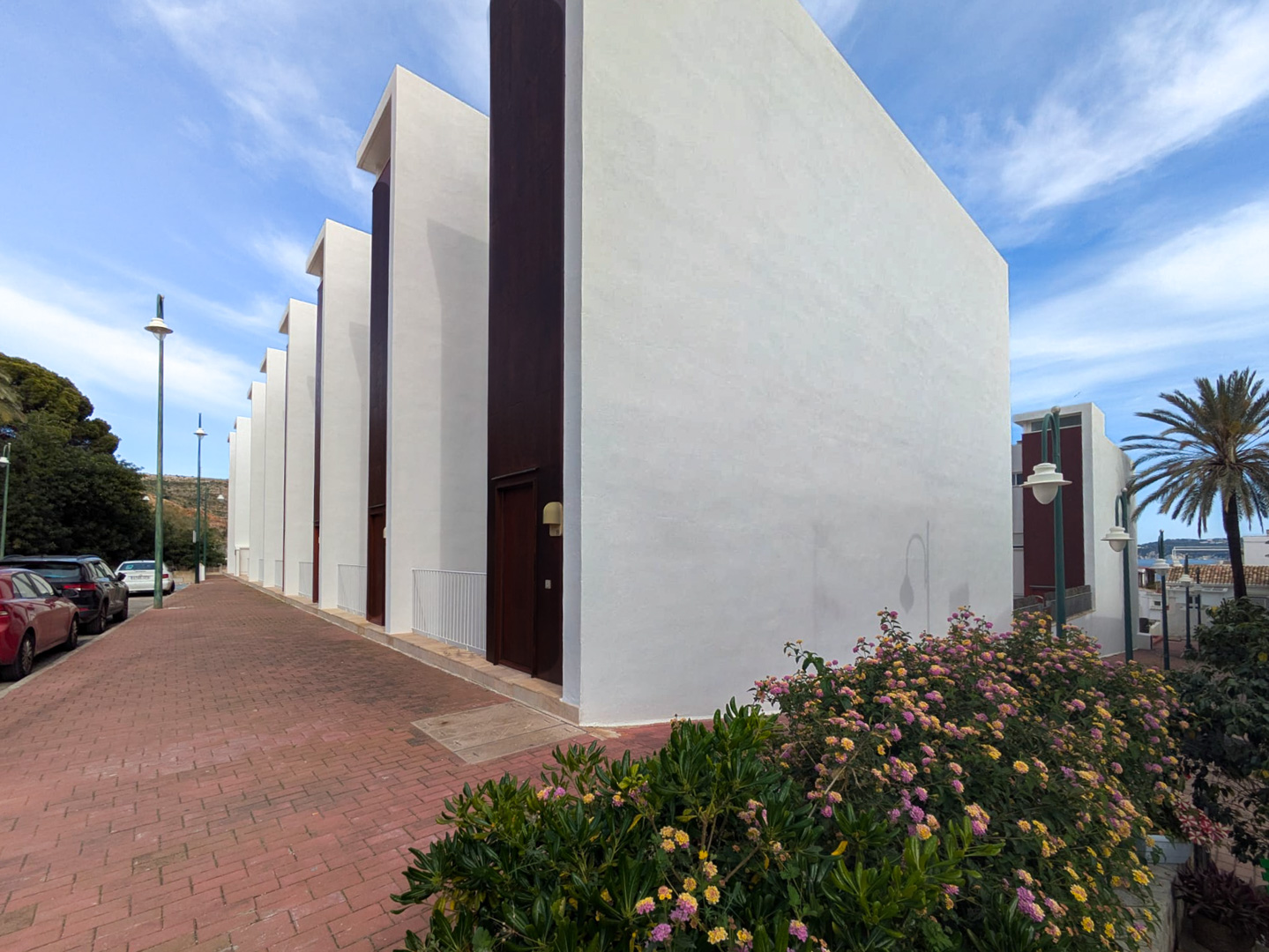 Façade extérieure au design contemporain avec des murs blancs et des panneaux verticaux sombres, bordée d'un trottoir pavé et de fleurs colorées.