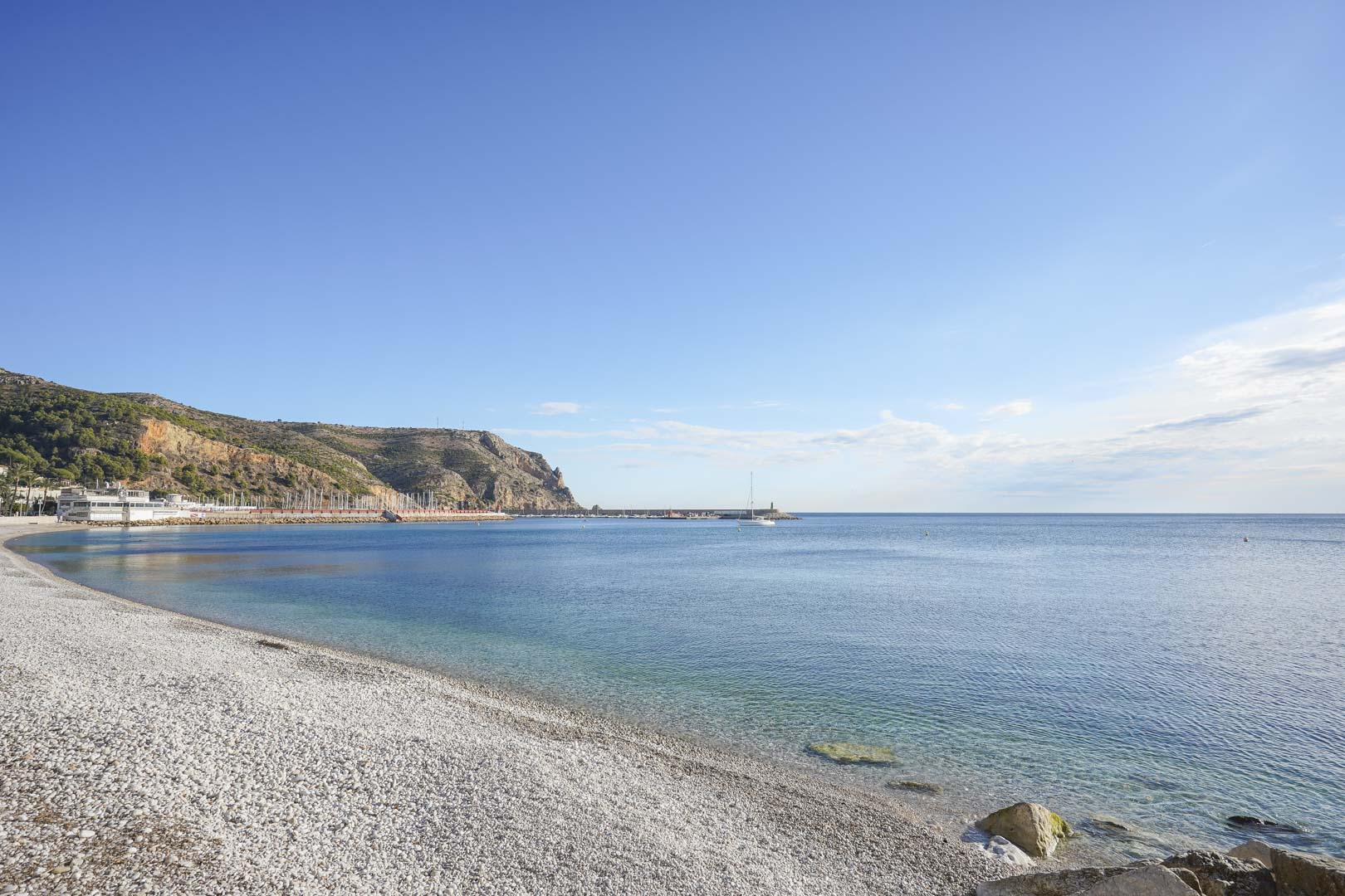 Vue de la plage de galets à Jávea, offrant des eaux cristallines et une vue sur le port de plaisance au pied du Cabo de San Antonio.