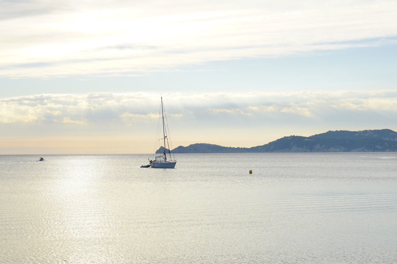 Vue sur la mer Méditerranée près du port de Jávea, avec un voilier sur des eaux calmes sous une lumière naturelle douce et la côte en arrière-plan.