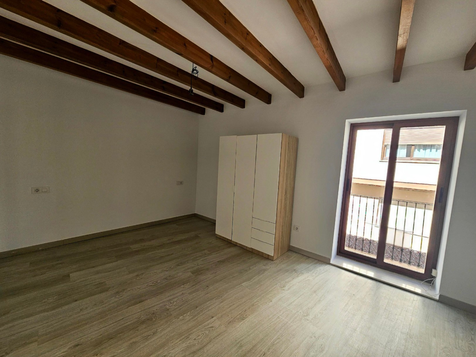 Chambre lumineuse avec poutres en bois apparentes, sol stratifié clair et accès direct à un balcon privé par une porte-fenêtre à double vitrage.