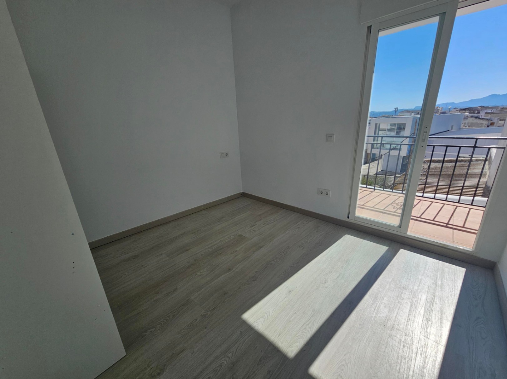 Chambre lumineuse avec sol en stratifié bois clair, murs blancs et porte vitrée coulissante menant à un balcon privé offrant une vue dégagée sur le village et les montagnes.