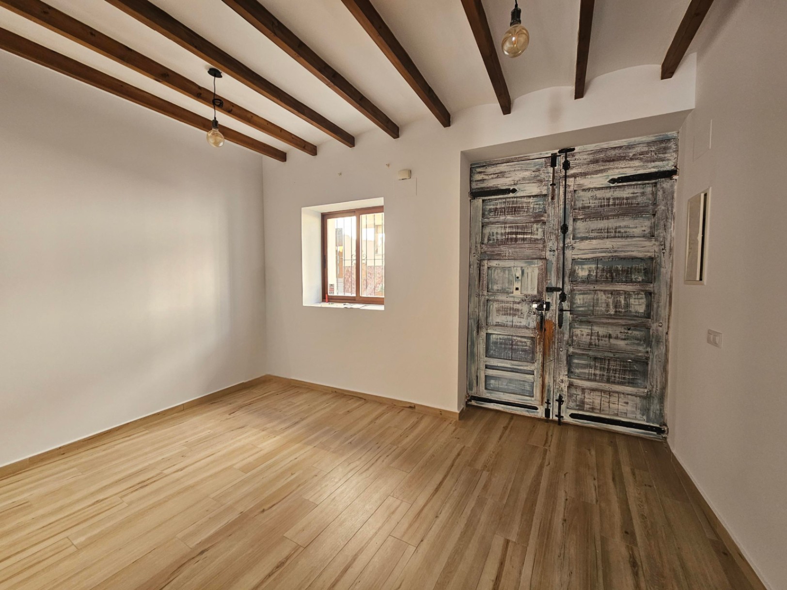 Espace intérieur avec poutres apparentes en bois sombre, sol clair et une grande porte double rustique au fini patiné avec des ferrures en fer traditionnelles.