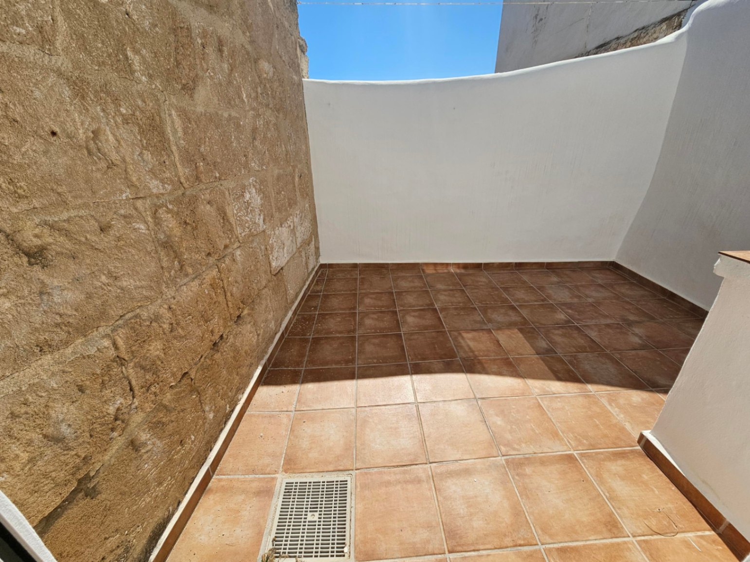 Patio extérieur privé avec sol en carreaux de terre cuite, mur en pierre naturelle d'origine et parois blanches sous un ciel dégagé.