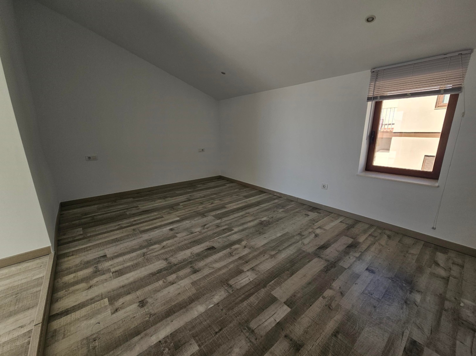 Pièce avec plafond mansardé, sol stratifié effet bois et spots encastrés. Fenêtre à cadre en bois avec store apportant une lumière naturelle.