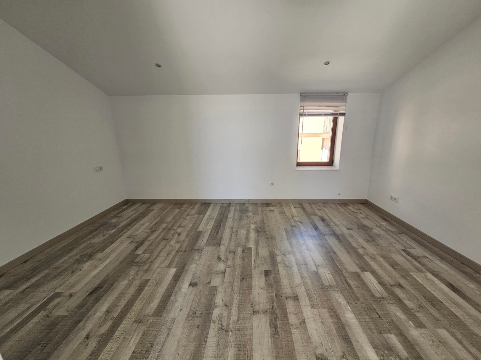 Pièce mansardée lumineuse avec sol en stratifié gris, murs blancs et spots encastrés au plafond. Une fenêtre à cadre en bois avec store complète cet espace rénové.