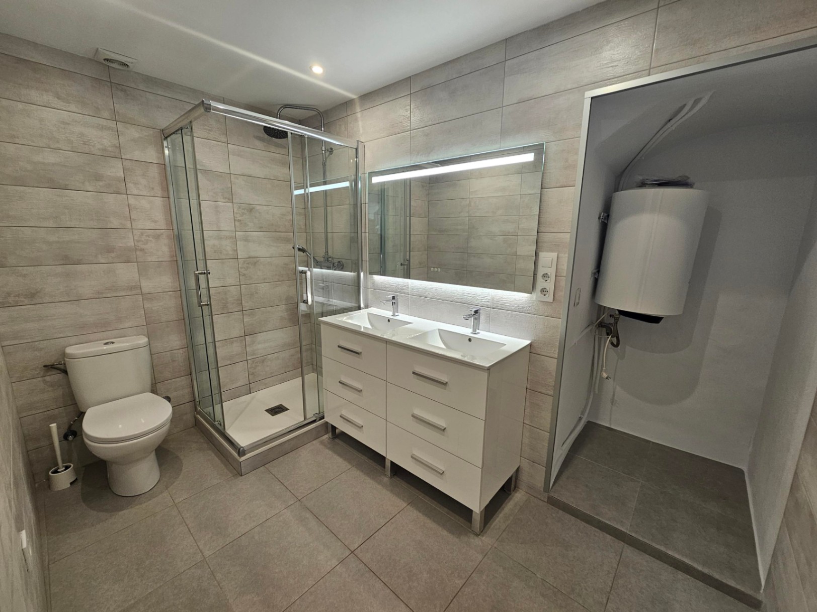 Salle de bain moderne avec double vasque, miroir LED et douche vitrée. Comprend un carrelage céramique gris et un espace technique pour le chauffe-eau électrique.
