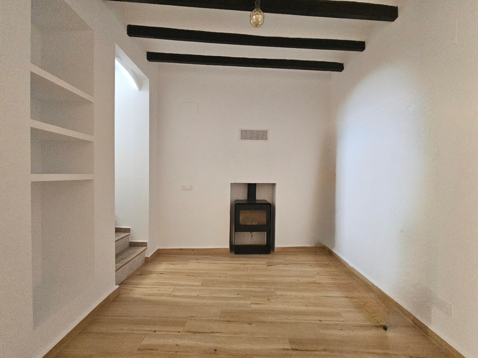 Salon avec poutres apparentes sombres, étagères intégrées et poêle à bois encastré. Le sol en bois clair et les murs blancs créent une atmosphère épurée.
