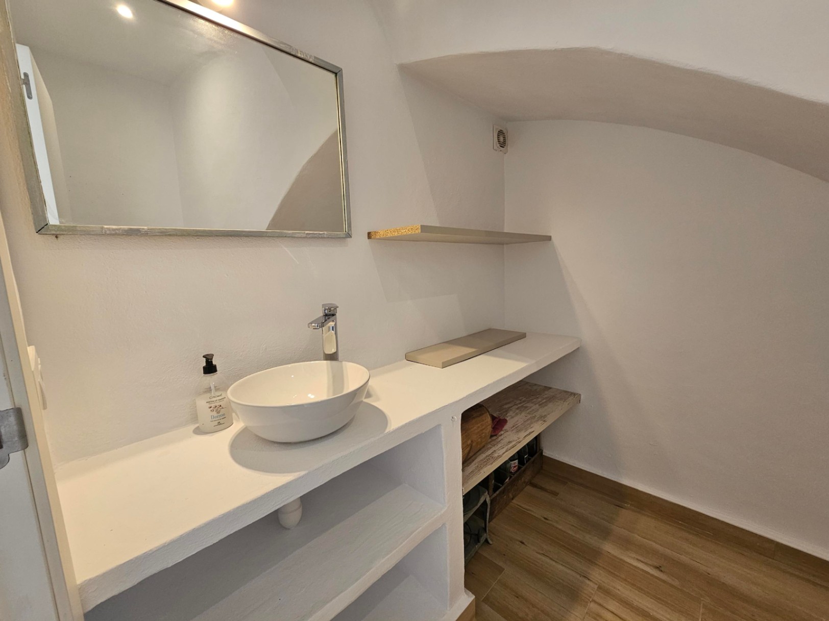 Toilettes d'invités avec vasque à poser sur plan de travail en maçonnerie blanche, étagères intégrées, sol imitation bois et plafond mansardé.