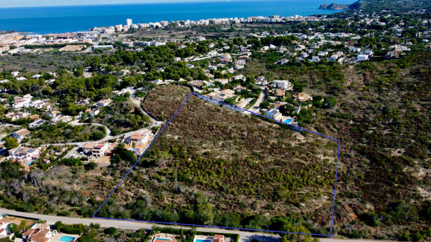 Vue aérienne du terrain de 17 272 m² à Jávea, délimité en bleu, entouré de végétation méditerranéenne avec une vue dégagée sur la côte et la mer.