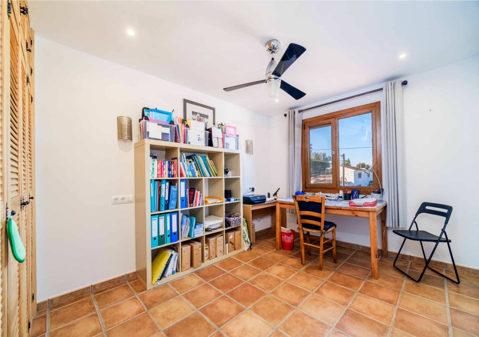 Bureau fonctionnel avec sol en terre cuite, étagère modulaire et bureau en bois. Équipé d'un ventilateur de plafond, de spots encastrés et de placards intégrés aux portes persiennes.