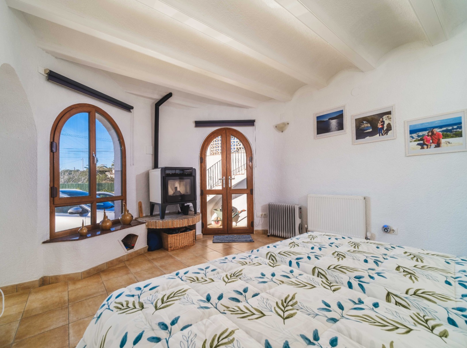 Chambre avec plafond à poutres blanches, poêle à bois sur socle en brique et menuiseries cintrées en bois offrant un accès vers l'extérieur.