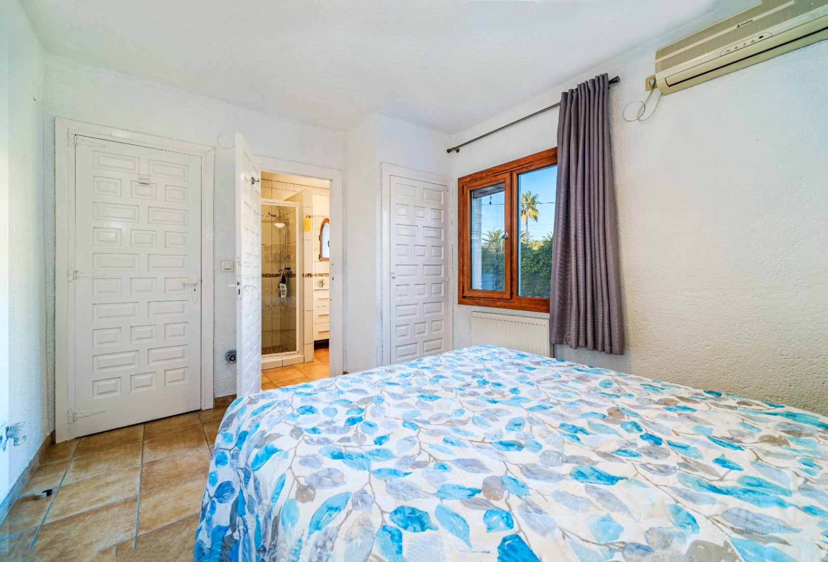 Chambre avec salle de bain attenante, portes blanches à panneaux, sol en céramique et fenêtre en bois. Équipée d'une climatisation et d'un radiateur.