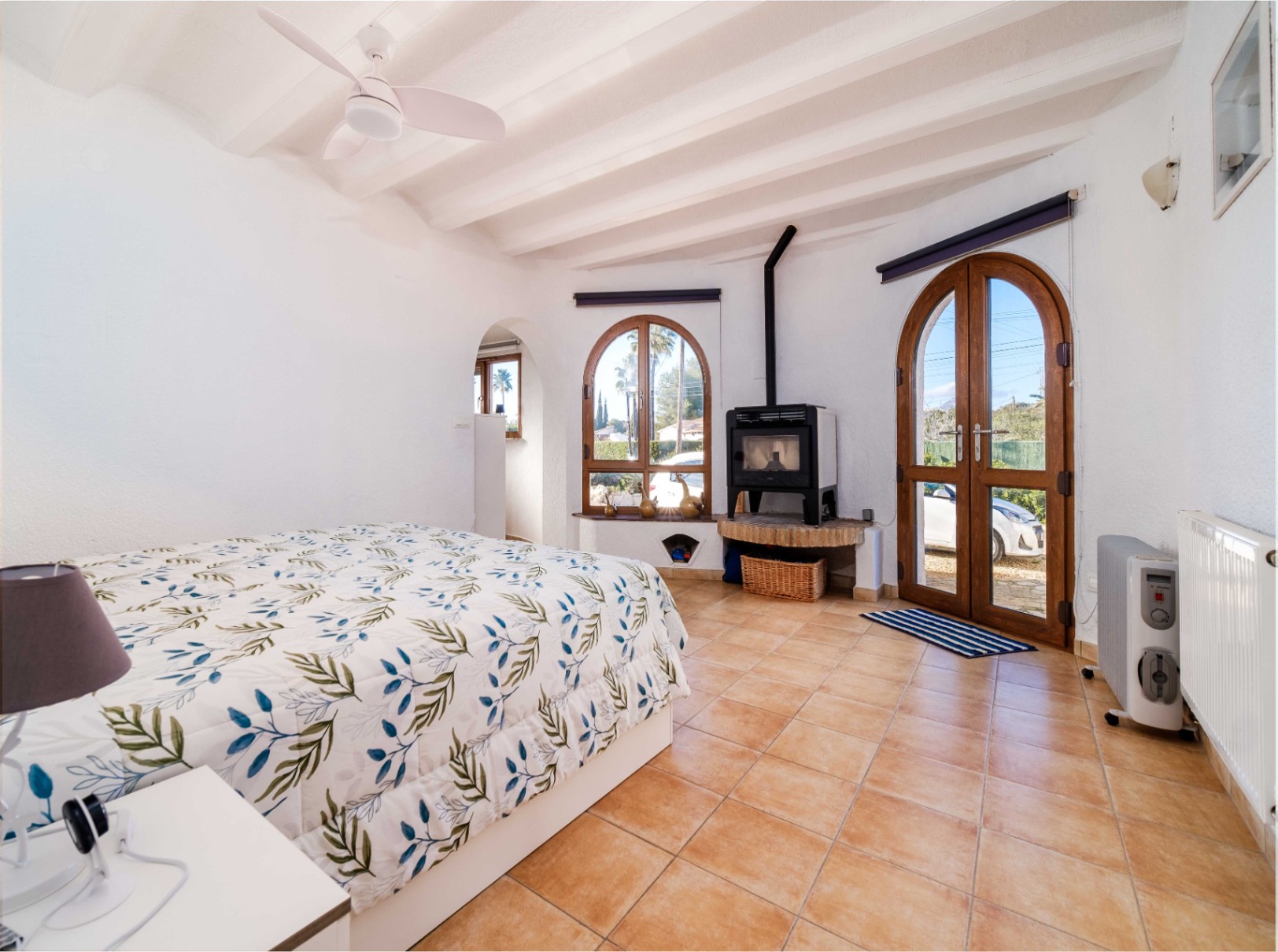 Chambre lumineuse avec plafond à poutres blanches, sol en terre cuite et poêle à bois. Des portes cintrées en bois offrent un accès direct à l'extérieur.