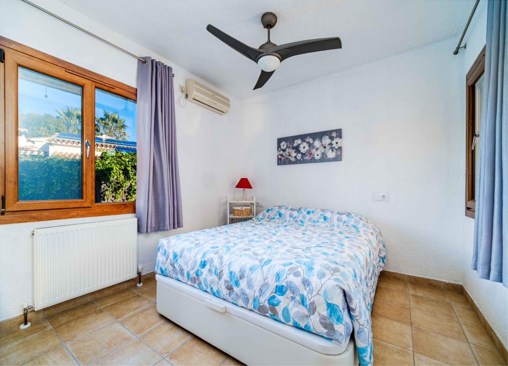 Chambre lumineuse avec sol en grès, ventilateur de plafond, climatisation réversible et fenêtres en bois offrant une vue sur le jardin.