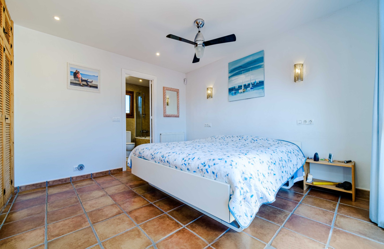 Chambre lumineuse avec sol en terre cuite, ventilateur de plafond et salle de bain attenante. Elle dispose de placards intégrés en bois et d'un éclairage soigné.