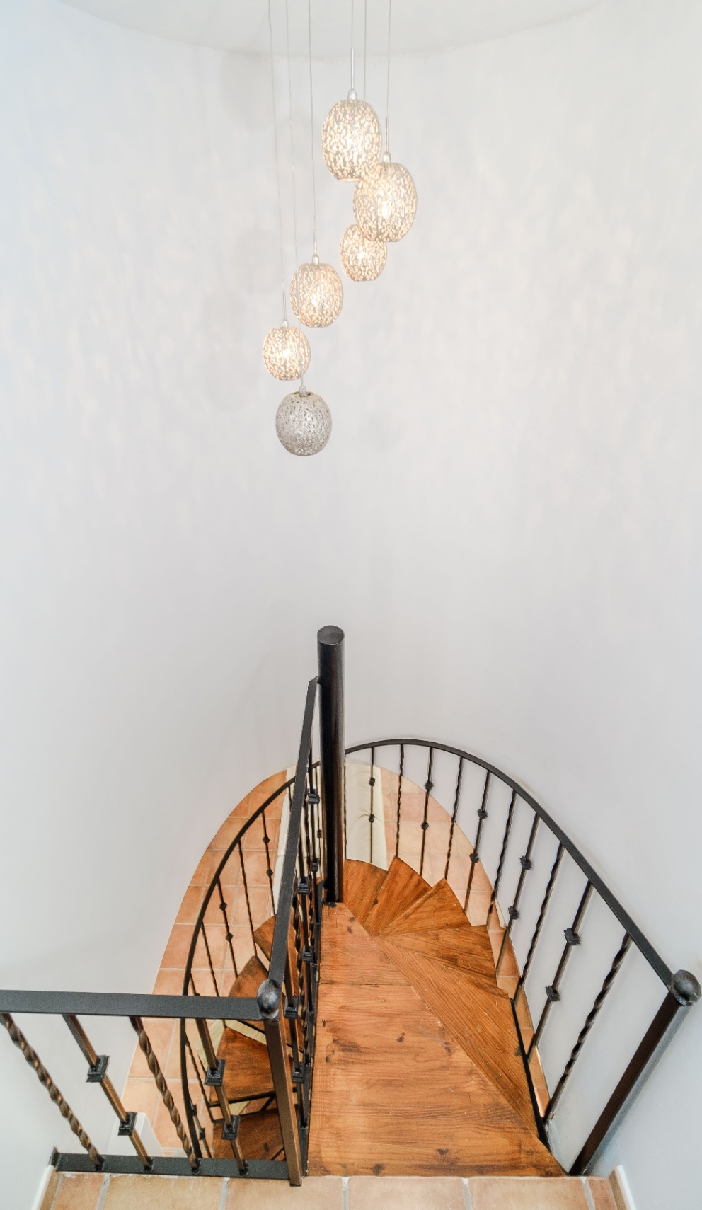 Escalier en colimaçon avec des marches en bois massif et une rampe en fer forgé noir, éclairé par une série de suspensions sphériques ajourées.