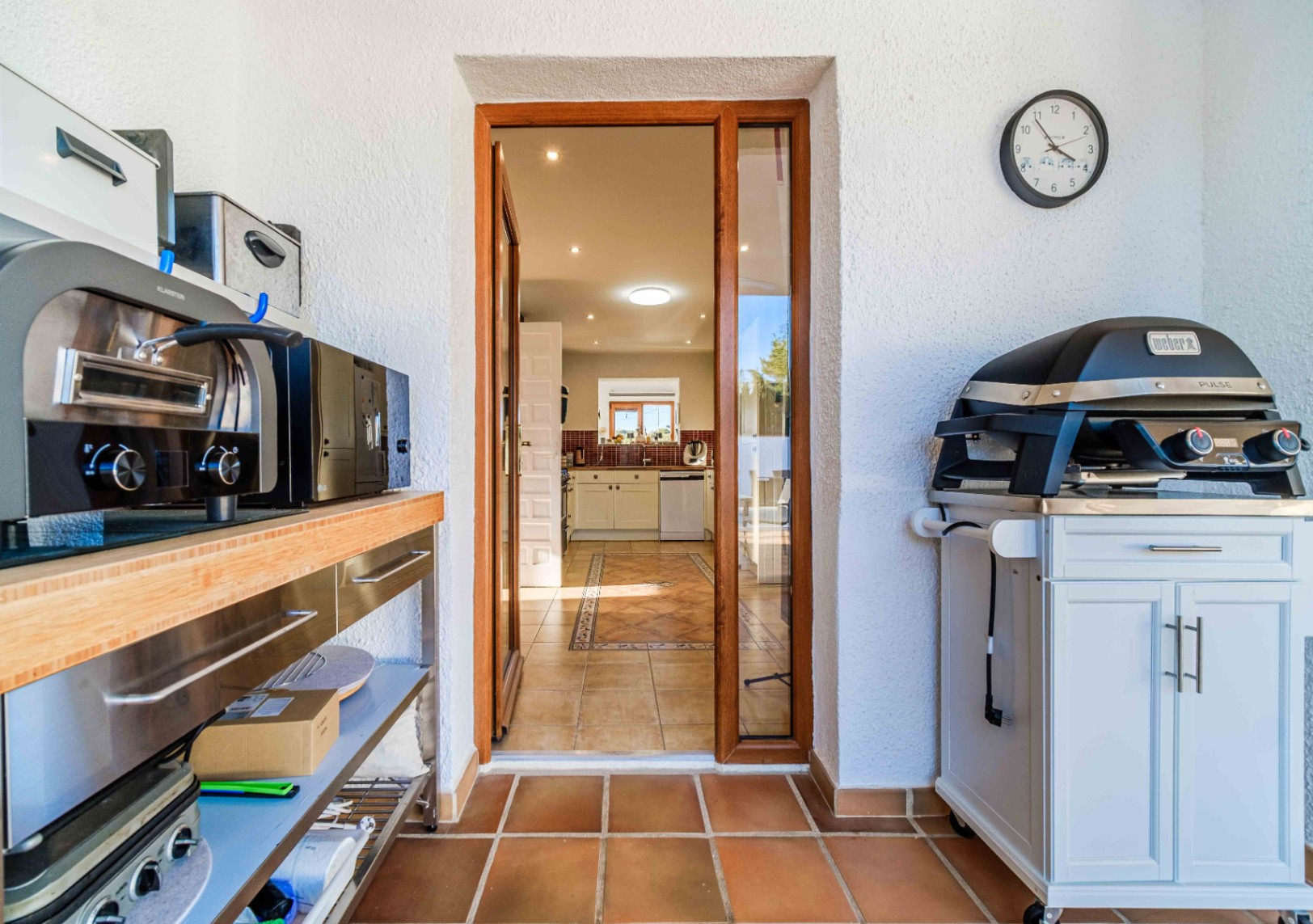 Espace cuisine extérieure avec barbecue électrique Weber et plan de travail en inox, offrant un accès direct à la cuisine intérieure par une porte vitrée.