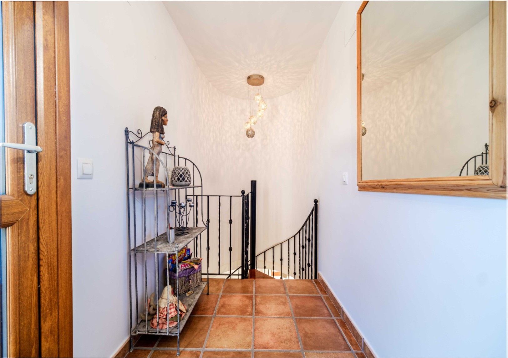 Palier avec sol en terre cuite, rampe d'escalier en fer forgé et luminaire suspendu créant des jeux d'ombre sur les murs blancs.