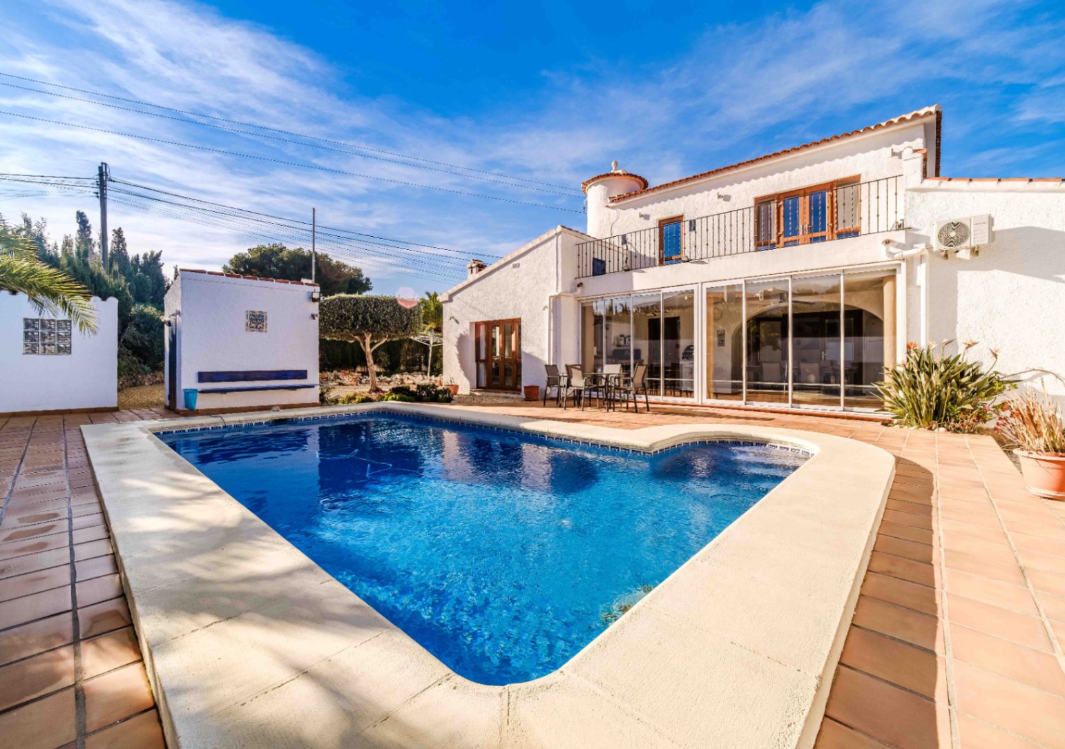 Piscine entourée d'une terrasse en terre cuite, offrant une vue sur la façade blanche de la villa et sa naya vitrée sous un ciel azur.