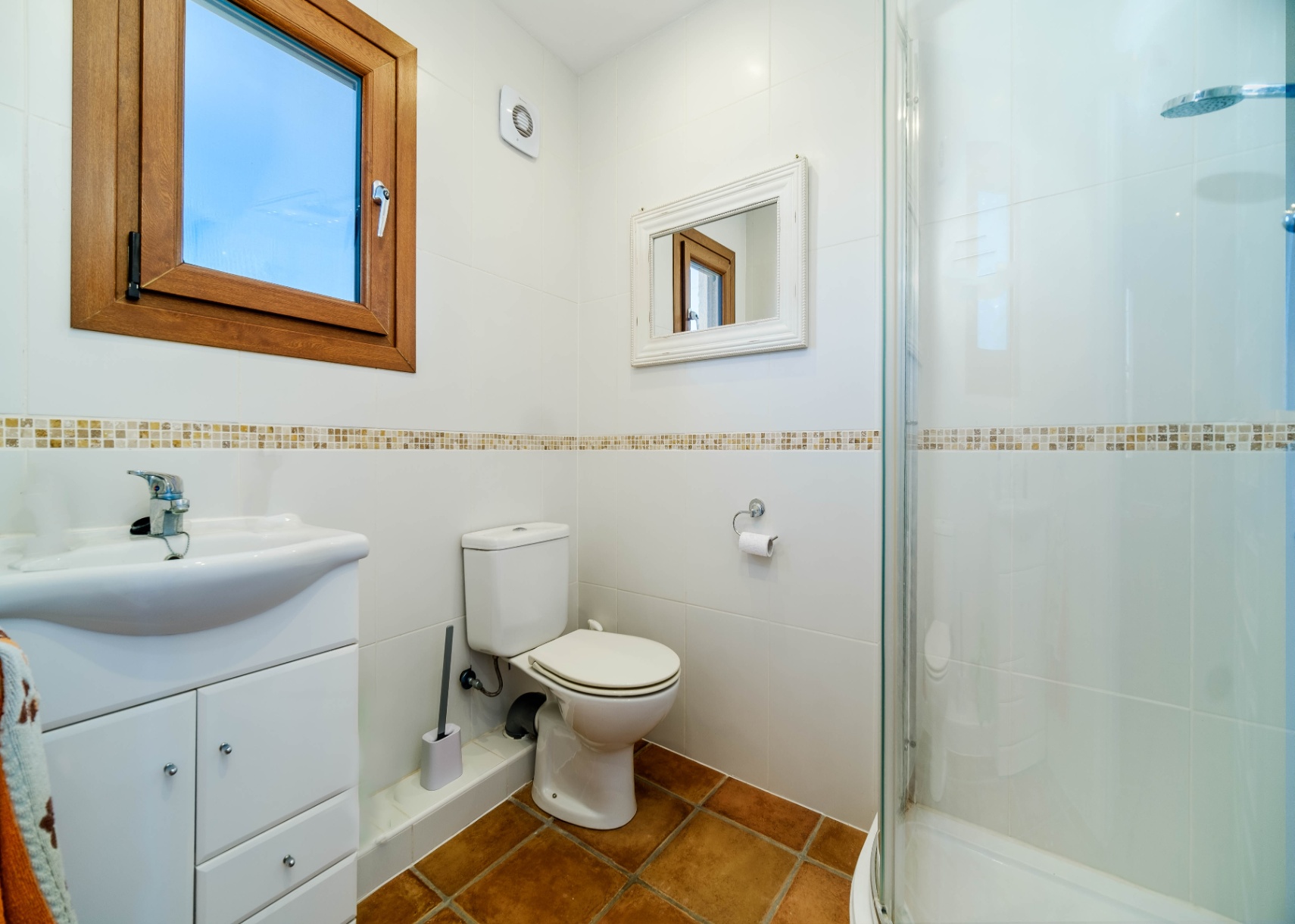 Salle de bain avec murs carrelés blancs, frise en mosaïque et sol en terre cuite. Elle dispose d'une douche vitrée, d'un meuble vasque et d'une fenêtre en bois.