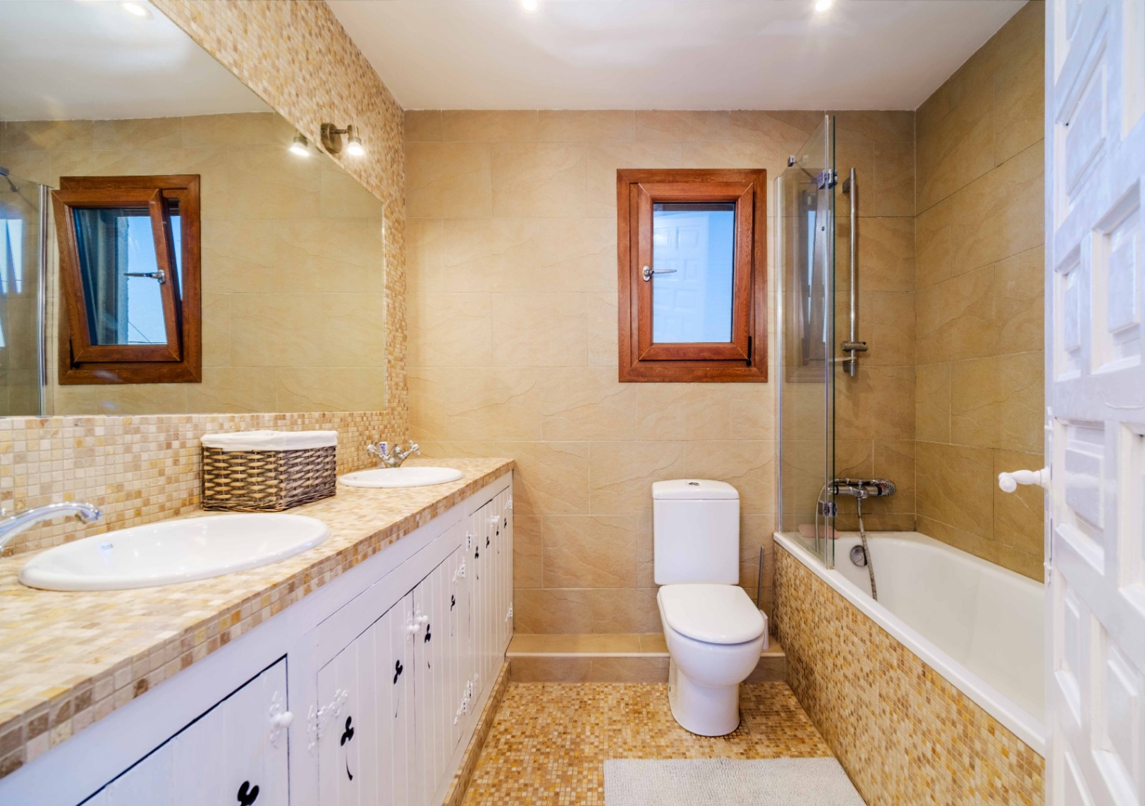 Salle de bain complète avec double vasque sur plan de travail en mosaïque, meubles blancs de style rustique et baignoire avec paroi en verre et carrelage aux tons terre.