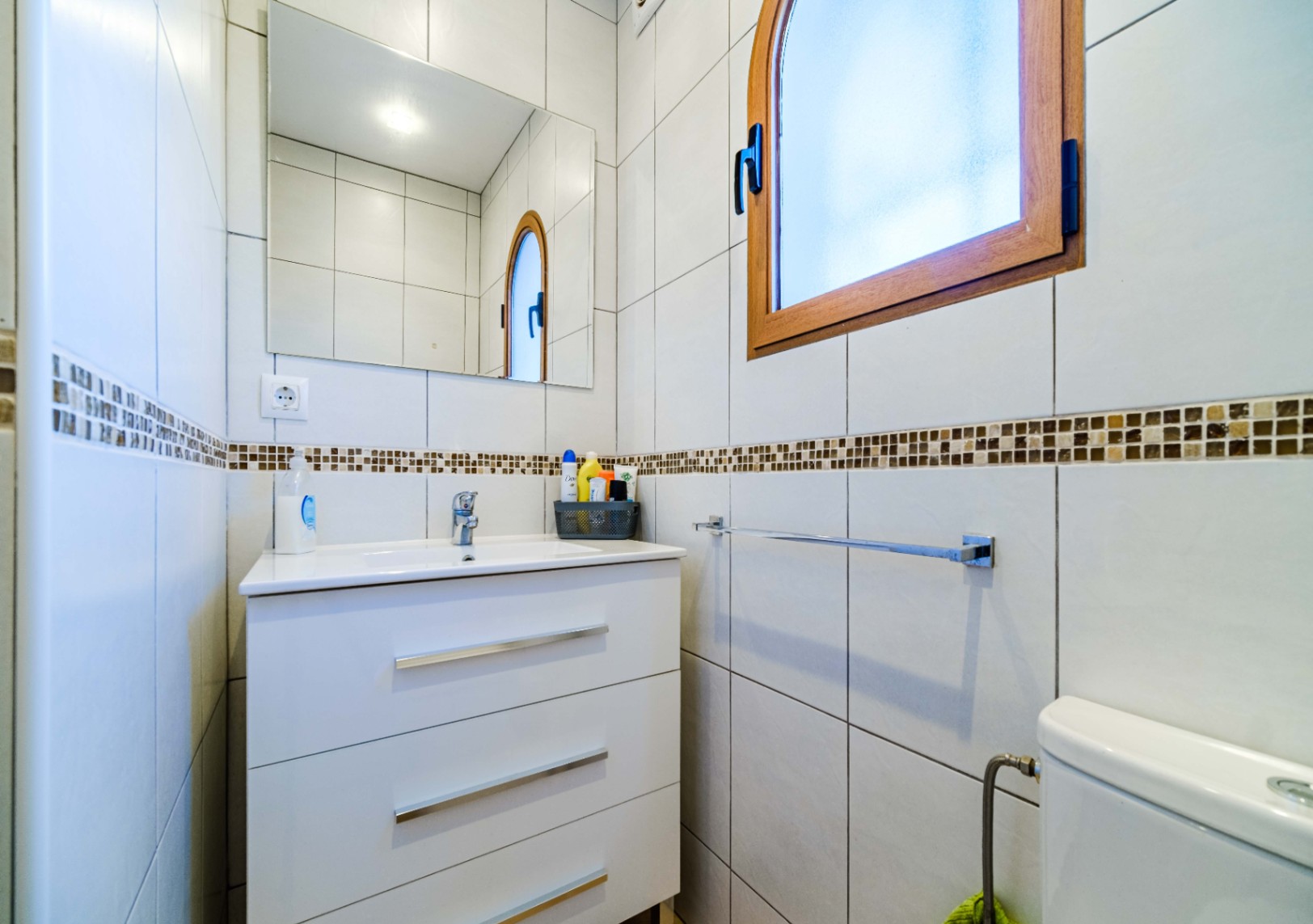 Salle de bain fonctionnelle avec meuble vasque blanc à trois tiroirs, miroir rectangulaire et fenêtre cintrée en bois. Murs carrelés avec une frise en mosaïque.