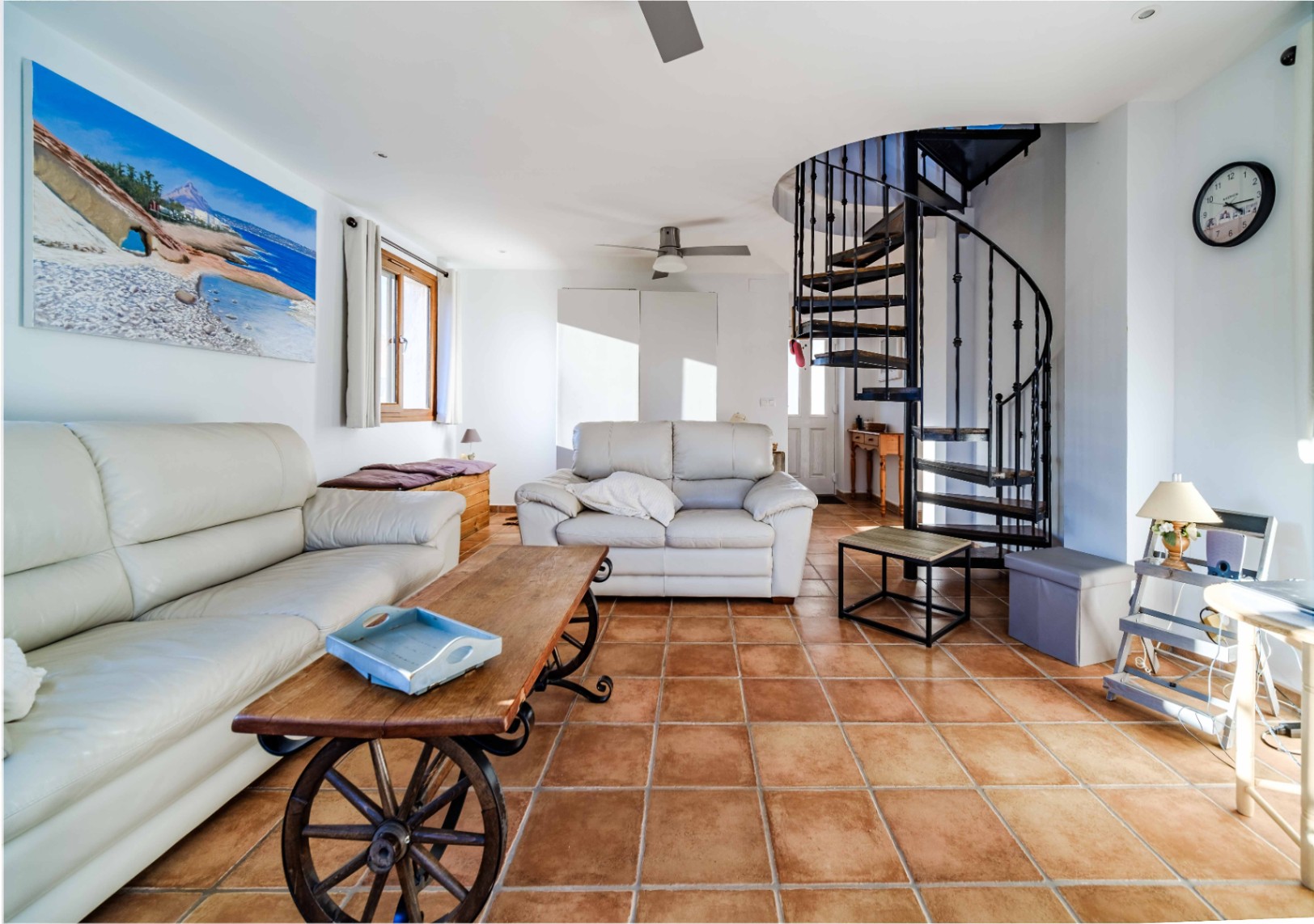 Salon avec sol en terre cuite, canapés en cuir blanc et table basse originale à roues. Un escalier en colimaçon en métal noir complète cet espace de vie fonctionnel.