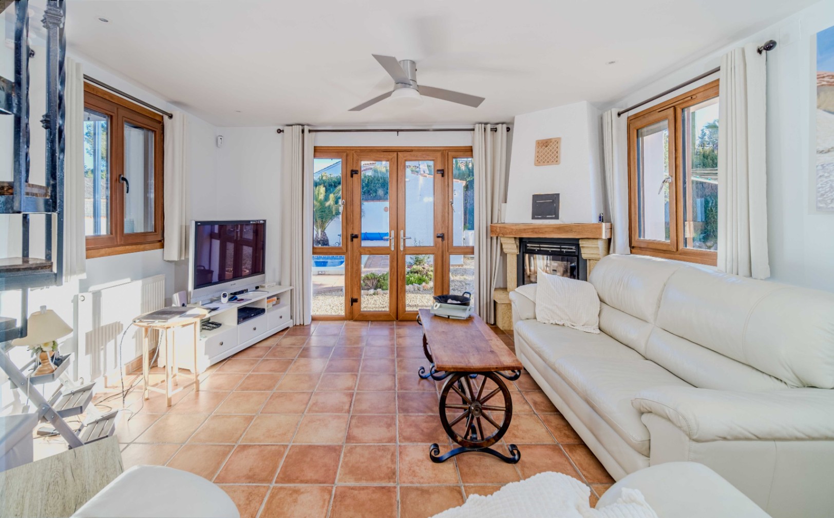 Salon lumineux avec sol en terre cuite, cheminée rustique et canapé en cuir blanc. Les portes-fenêtres en bois offrent un accès direct à la piscine.