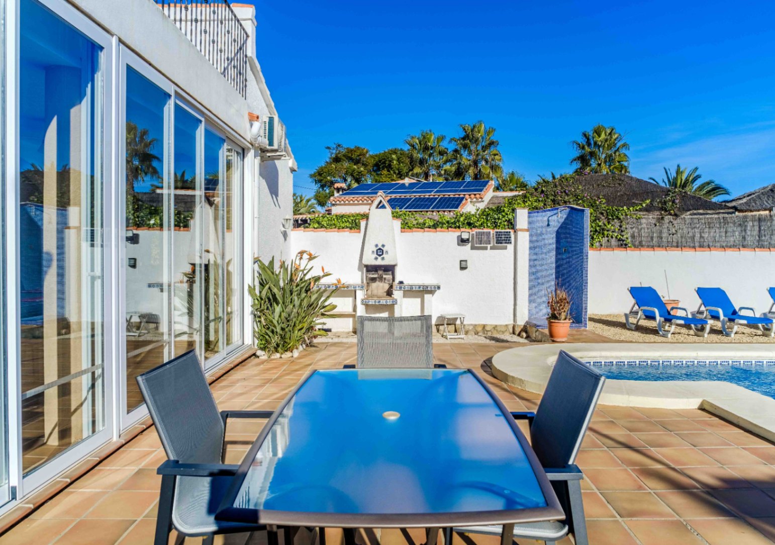 Terrasse extérieure avec coin repas, barbecue intégré et douche en mosaïque bleue. Les grandes baies vitrées et les panneaux solaires soulignent la modernité du lieu.