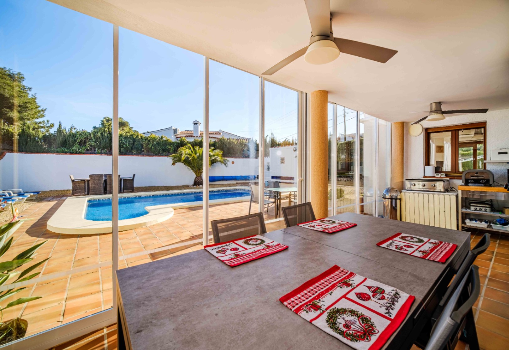Terrasse vitrée avec coin repas, ventilateurs de plafond et cuisine d'été, offrant une vue imprenable sur la piscine et les extérieurs.