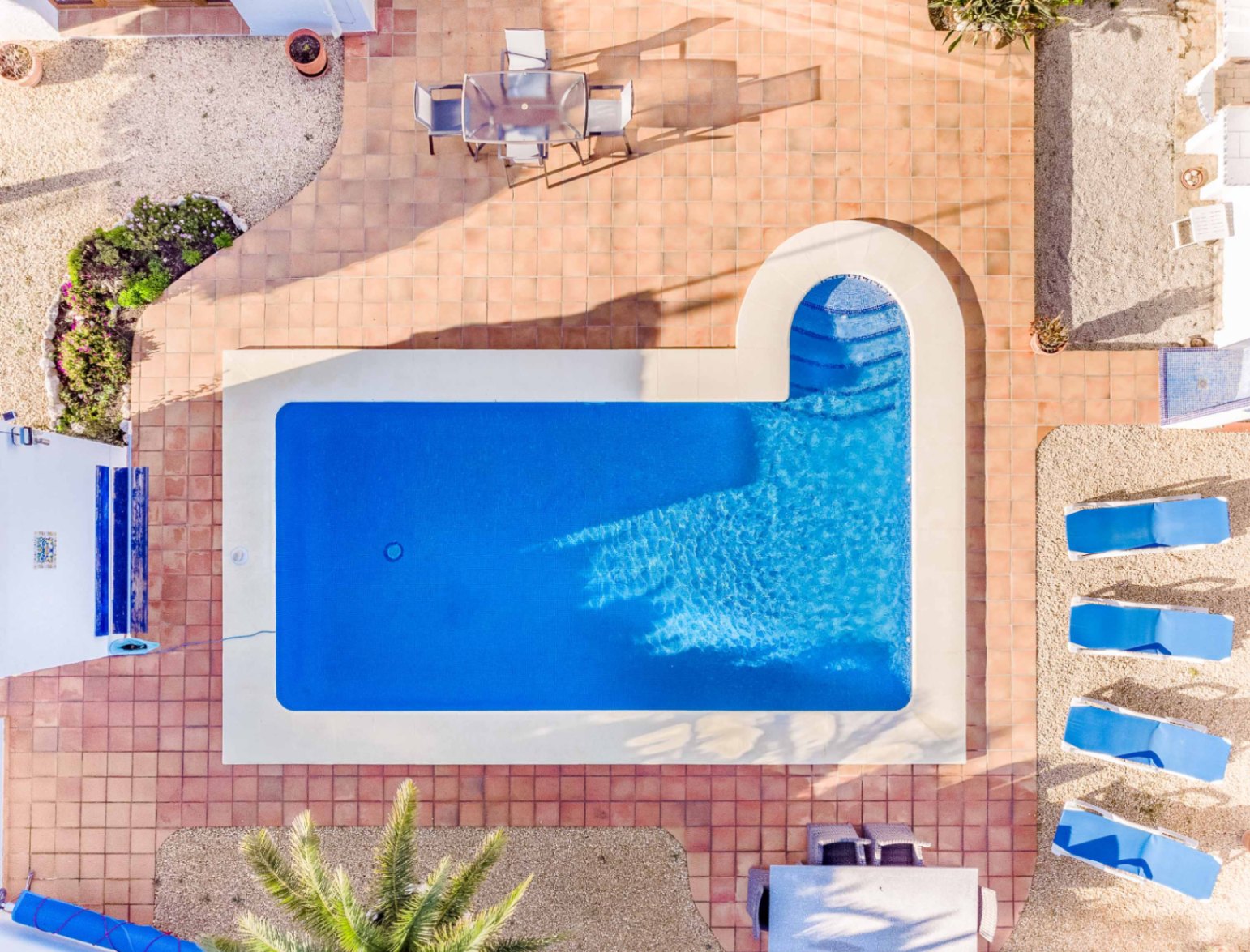 Vue aérienne de la piscine rectangulaire avec escalier romain, entourée d'une terrasse en terre cuite et d'un espace de détente avec bains de soleil bleus.