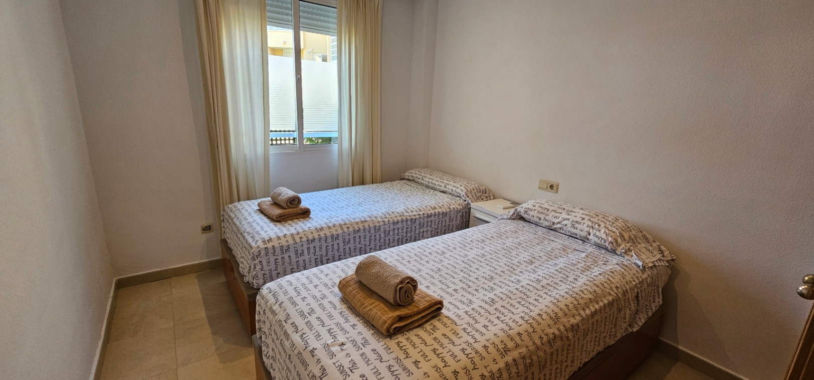 Chambre avec deux lits simples, sol en carrelage clair et fenêtre avec rideaux blancs apportant une luminosité naturelle.