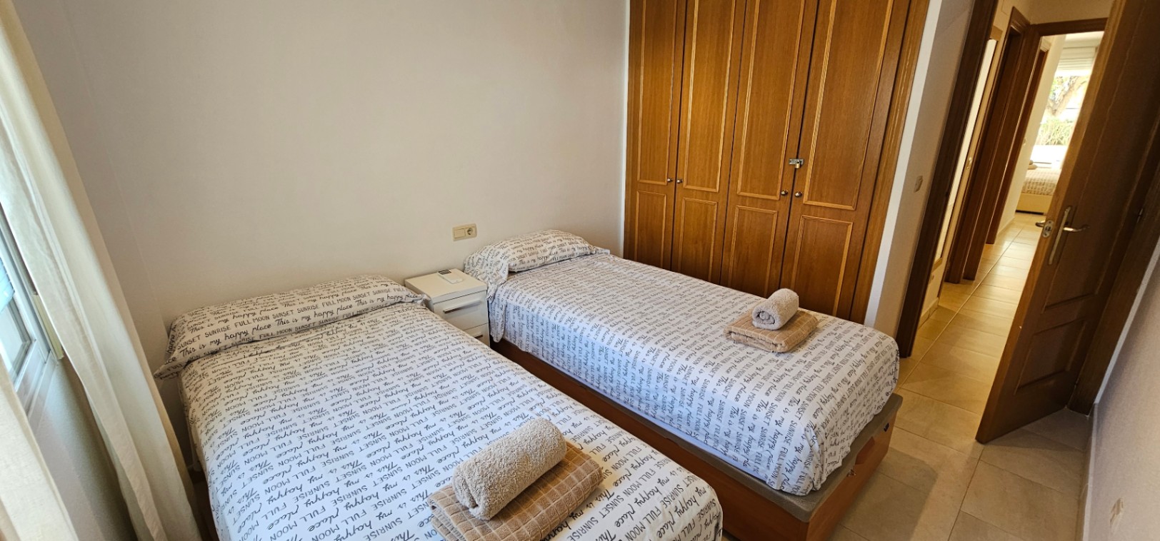 Chambre équipée de deux lits simples, d'un grand placard intégré en bois et d'un sol en carrelage clair, avec vue sur le couloir.