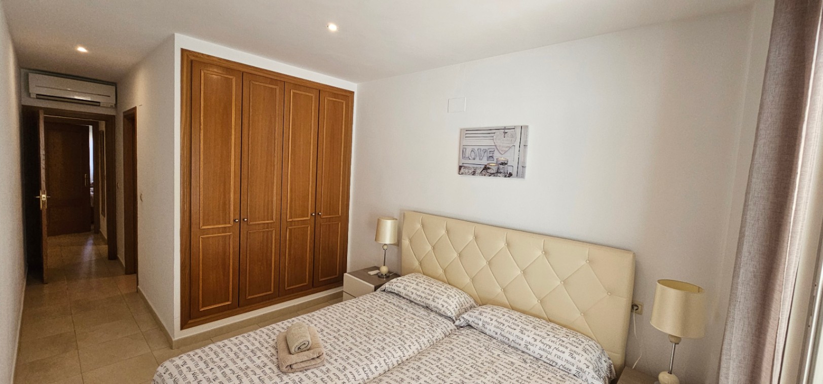 Chambre principale avec placards intégrés en bois, tête de lit capitonnée de couleur crème et climatisation installée au-dessus de la porte.