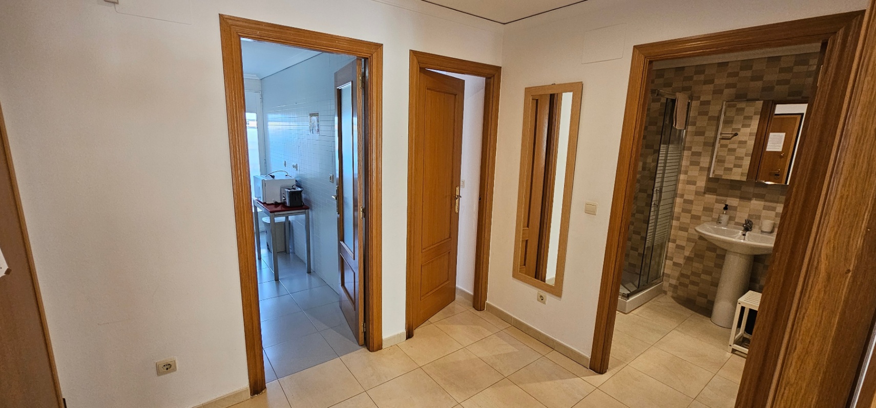 Couloir de distribution avec menuiseries en bois, desservant une cuisine carrelée et une salle de bain avec douche à l'italienne et faïence mosaïque.