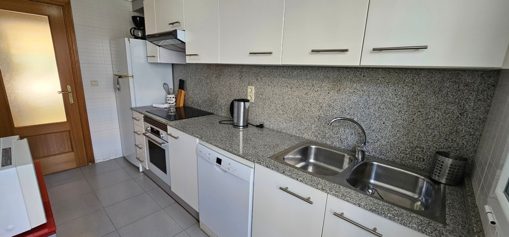 Cuisine fonctionnelle avec plan de travail et crédence en granit gris, meubles blancs, électroménagers intégrés et évier double en acier inoxydable.