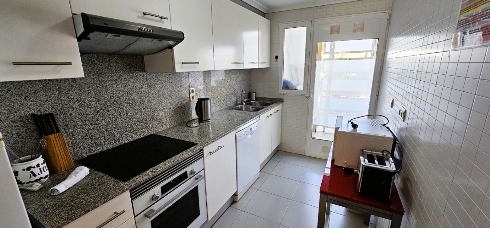 Cuisine indépendante avec plan de travail en granit, meubles blancs, four intégré, plaque vitrocéramique et porte vitrée menant vers l'extérieur.