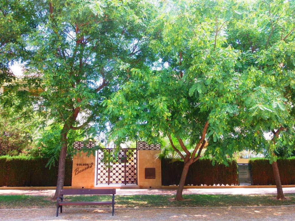 Entrée principale du complexe Golden Beach 2, avec portail en fer forgé, piliers en pierre et arbres matures ombrageant un banc de repos en bois.