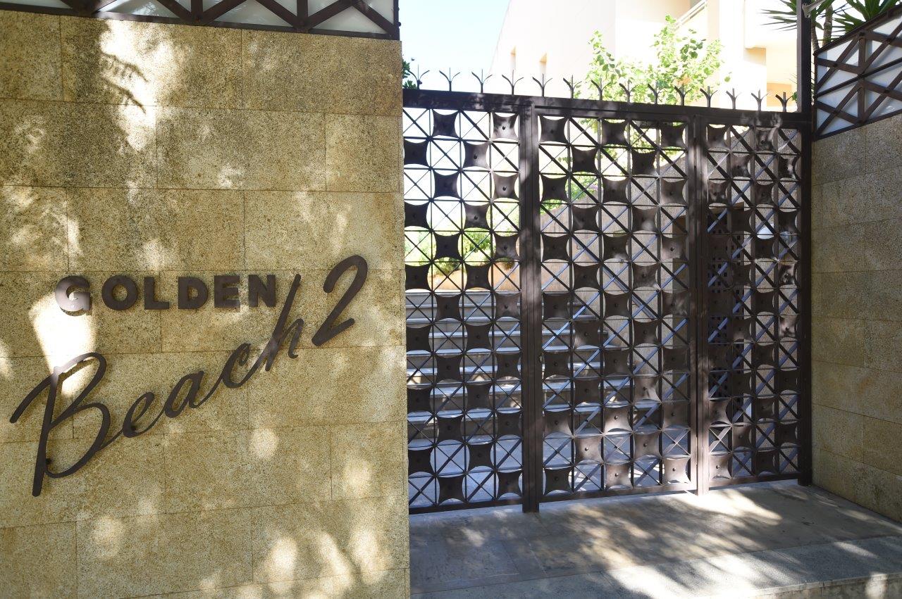 Entrée principale du complexe Golden Beach 2, avec un mur en pierre naturelle, une enseigne métallique et un portail décoratif en fer forgé.