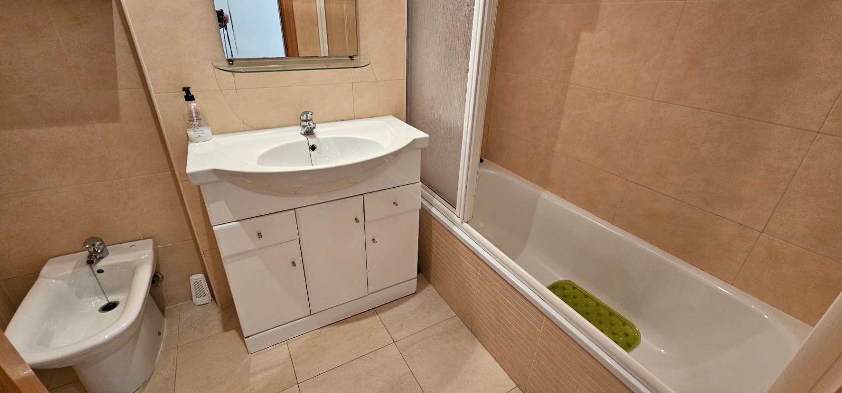 Salle de bain carrelée dans les tons crème, comprenant une baignoire avec paroi, un bidet et un meuble vasque blanc avec rangements.