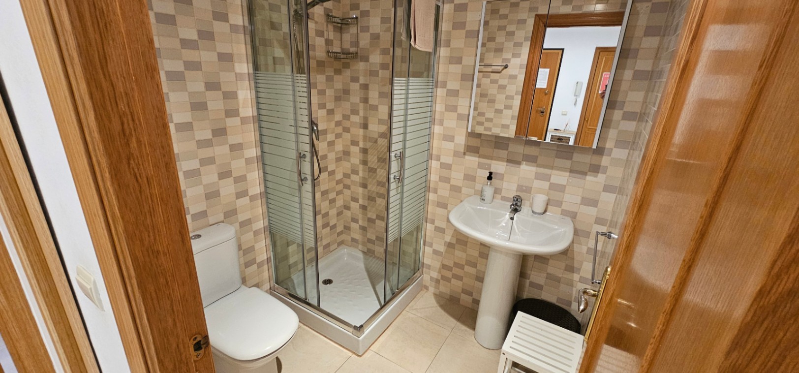 Salle de bain équipée d'une cabine de douche en angle, d'un lavabo sur colonne et de murs en mosaïque dans des tons naturels.