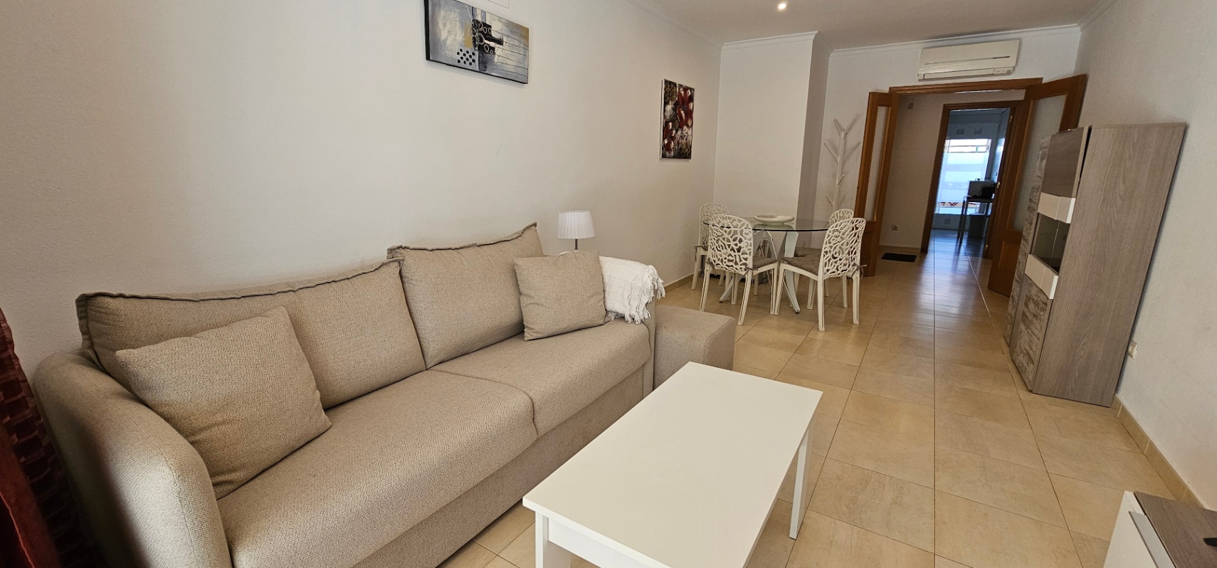 Salon-salle à manger avec canapé beige, table basse blanche et coin repas. La pièce dispose de sols en grès cérame et d'une climatisation murale installée.