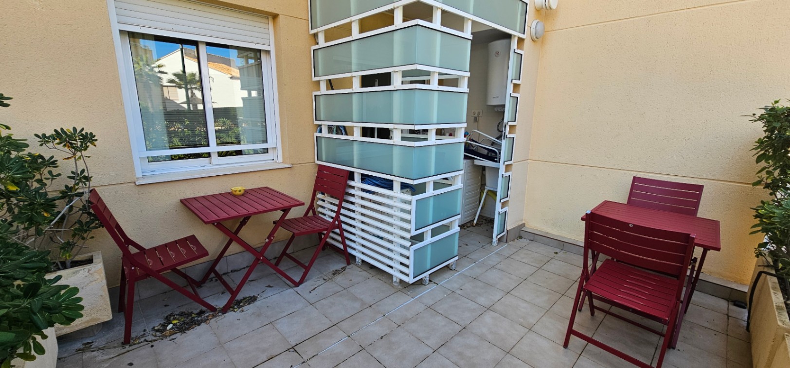 Terrasse privée avec carrelage au sol, mobilier de jardin bordeaux et un abri technique en lames blanches et verre dépoli pour le coin buanderie.