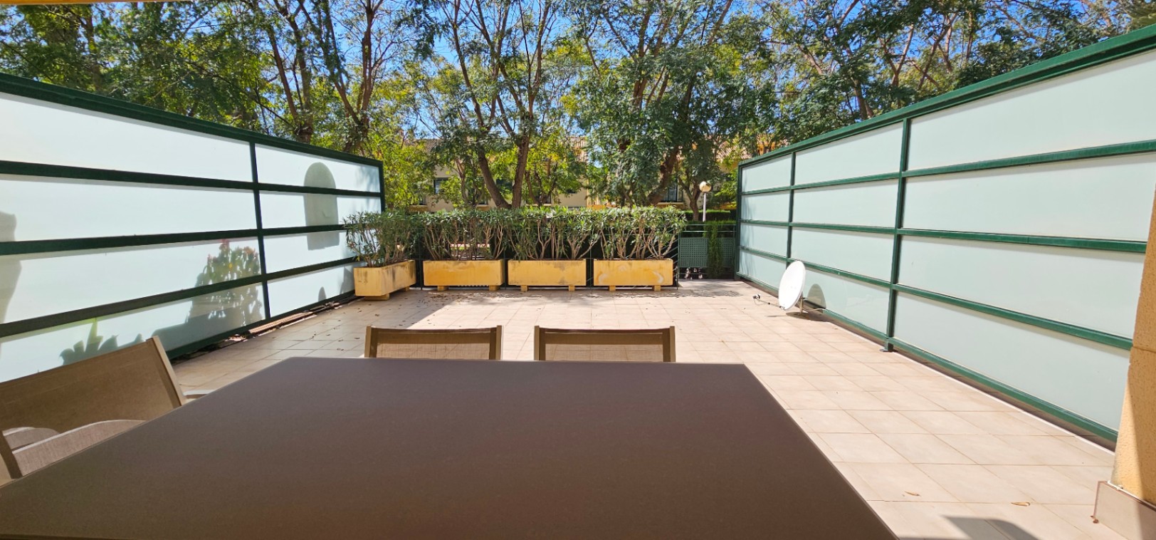 Terrasse privée avec revêtement en carrelage, parois latérales en verre dépoli pour l'intimité et jardinières bordées d'arbres matures environnants.