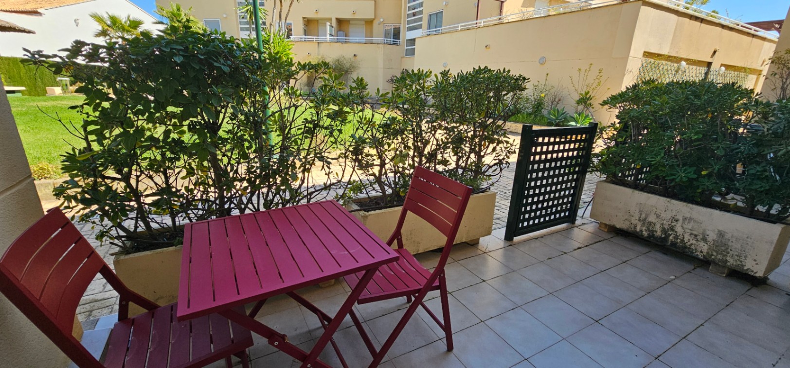 Terrasse privée carrelée avec mobilier en bois bordeaux et jardinières de haies, offrant un accès direct par portillon aux jardins de la résidence.