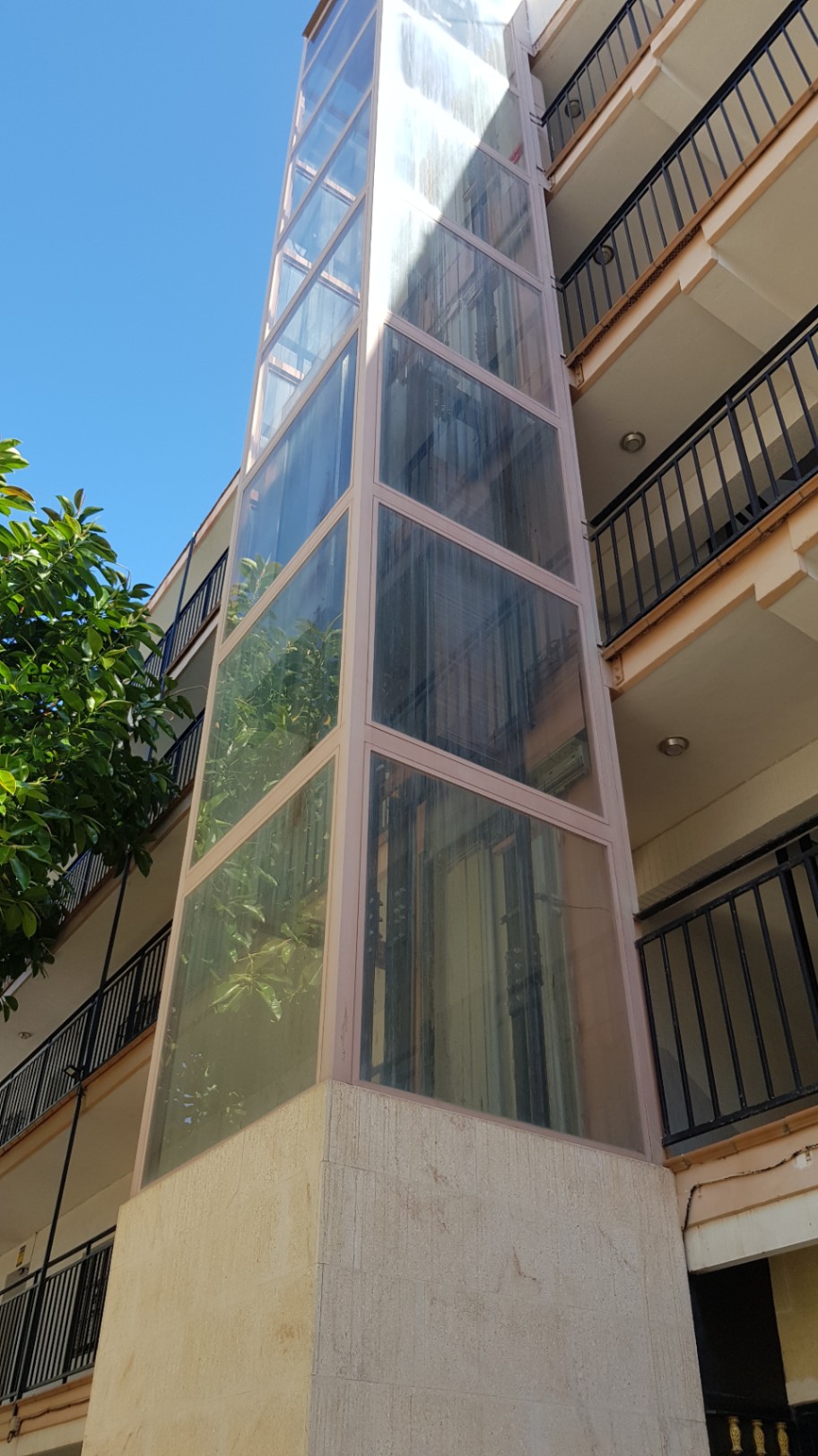 Ascenseur panoramique extérieur en verre avec base en pierre, intégré à la façade du bâtiment résidentiel et ses balcons aux garde-corps noirs.