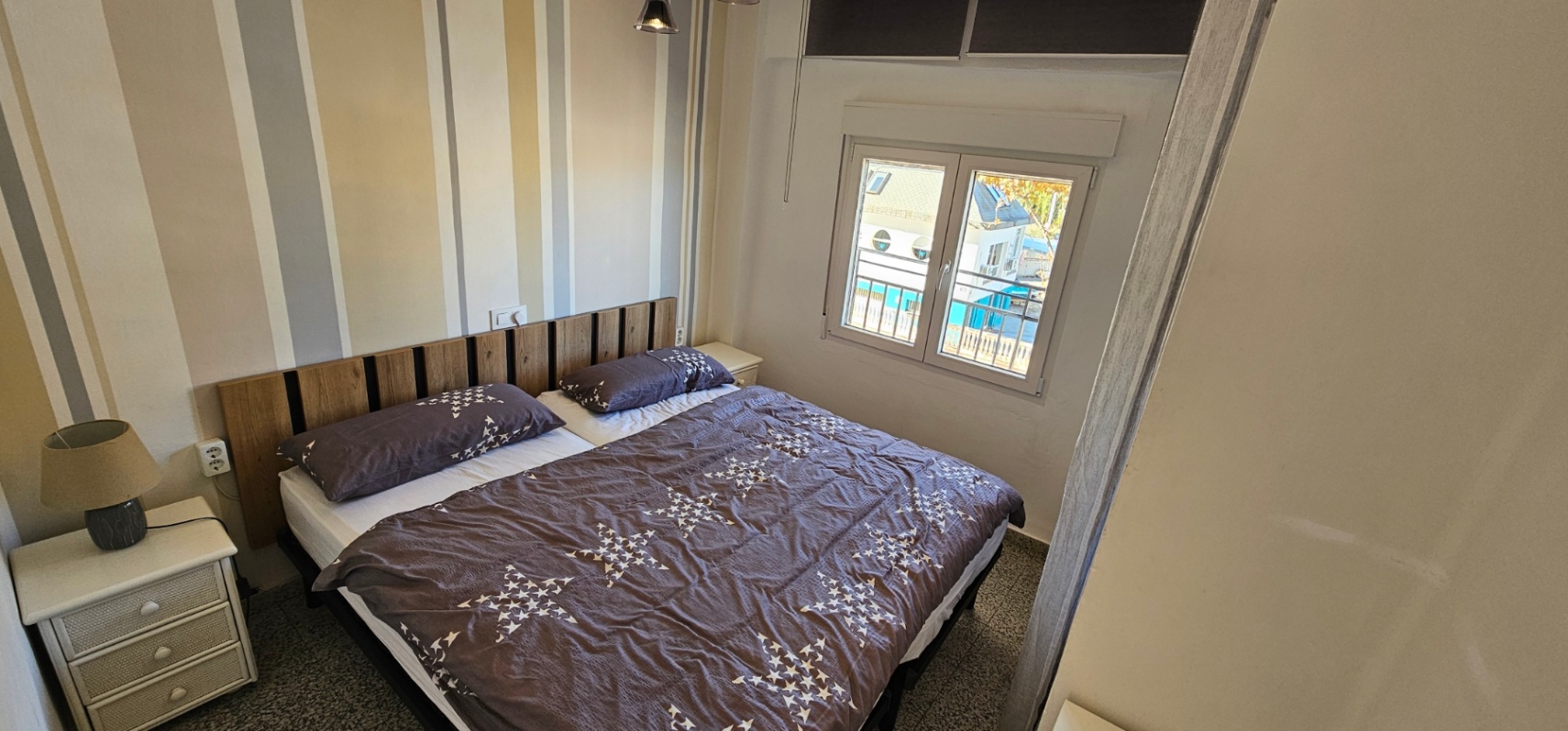 Chambre à coucher avec lit double, tête de lit en lattes de bois et papier peint rayé. Une fenêtre extérieure assure une luminosité naturelle constante.