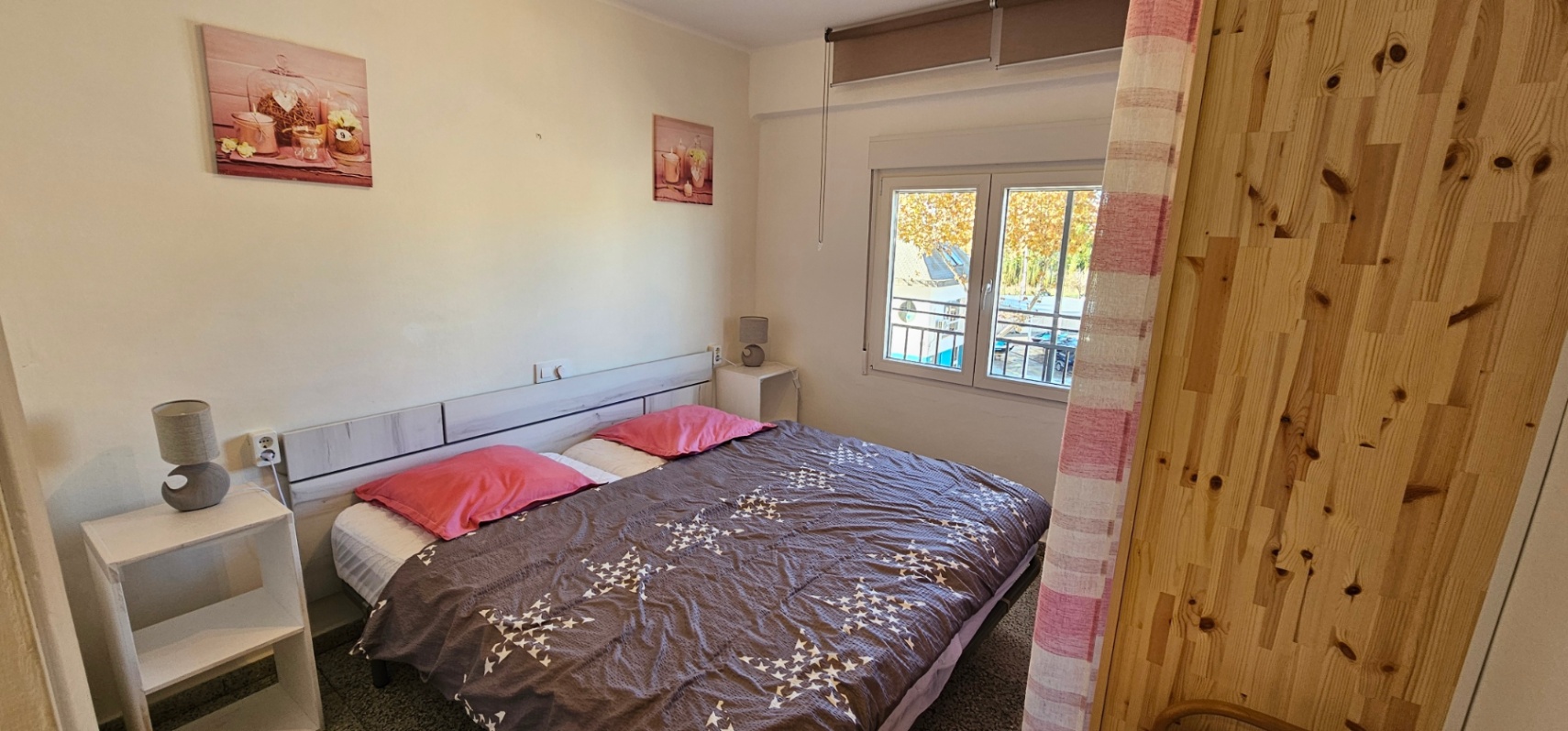 Chambre avec lit double, tête de lit en bois clair et tables de chevet blanches. Une fenêtre extérieure laisse entrer la lumière naturelle avec vue sur les environs.